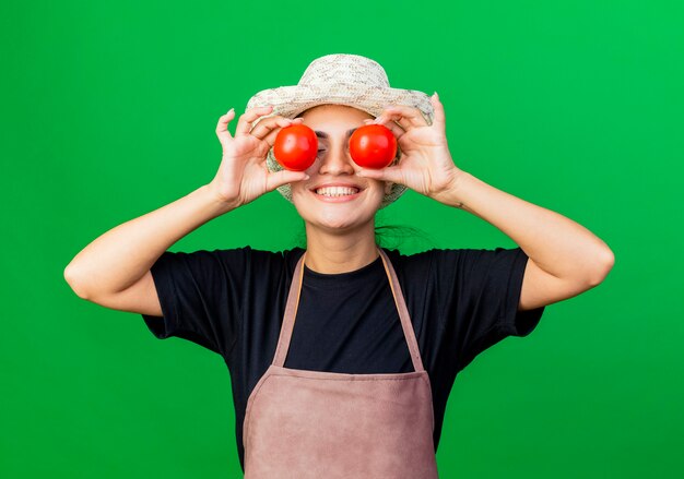 Молодая красивая женщина-садовник в фартуке и шляпе держит помидоры, закрывая глаза, улыбаясь