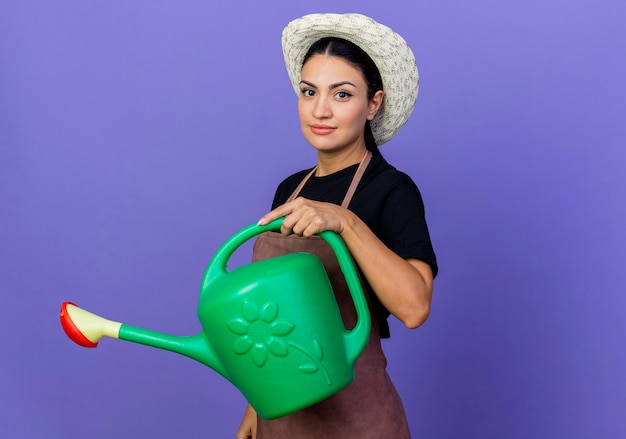 Giardiniere della giovane bella donna in grembiule e cappello che tiene annaffiatoio guardando davanti con faccia seria in piedi sopra la parete blu