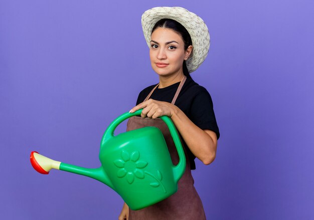 Young beautiful woman gardener in apron and hat holding watering can looking at front with serious face standing over blue wall