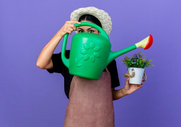 Young beautiful woman gardener in apron and hat holding watering can and potted plant standing over blue wall