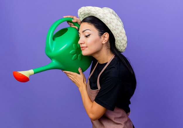 Young beautiful woman gardener in apron and hat holding watering can standing over blue wall