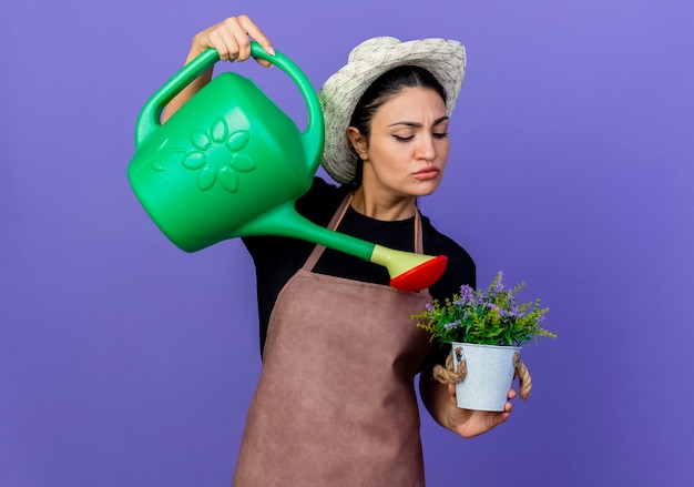 Молодая красивая женщина-садовник в фартуке и шляпе держит лейку, поливает горшечное растение, стоящее над синей стеной