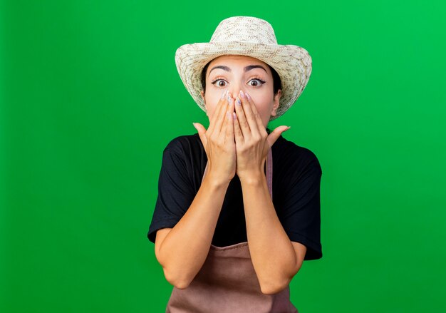 Young beautiful woman gardener in apron and hat looking at front covering mouth with hands being amazed and surprised standing over green wall