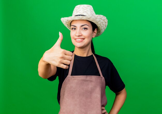 Молодая красивая женщина-садовник в фартуке и шляпе, глядя вперед, улыбаясь, показывает палец вверх, стоя над зеленой стеной