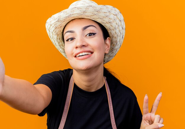 Молодая красивая женщина-садовник в фартуке и шляпе, глядя на переднюю улыбку, показывая знак v, стоящий над оранжевой стеной