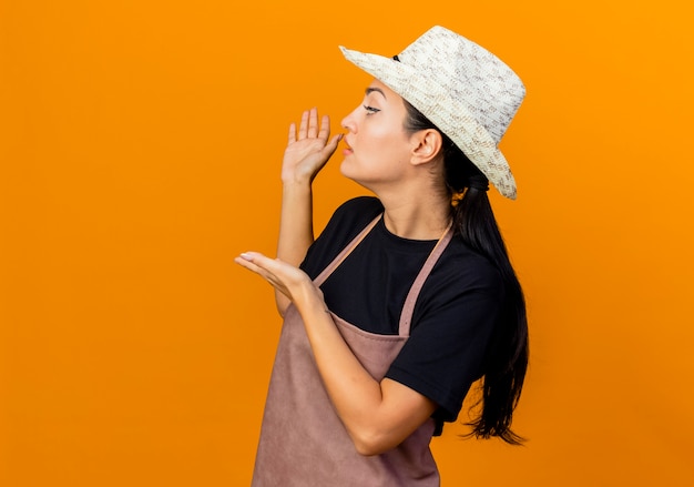 Young beautiful woman gardener in apron and hat presenting something with hands behind her standing over orange wall