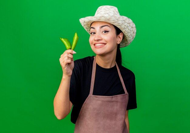 Молодая красивая женщина-садовник в фартуке и шляпе показывает сломанный зеленый перец чили, глядя вперед, улыбаясь со счастливым лицом, стоящим над зеленой стеной