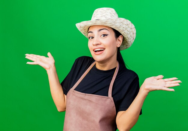 Young beautiful woman gardener in apron and hat smiling confused spreading arms to the sides 