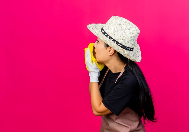 ピンクの壁の上に立っている手で誰かを呼び出すゴム手袋エプロンと帽子の若い美しい女性の庭師
