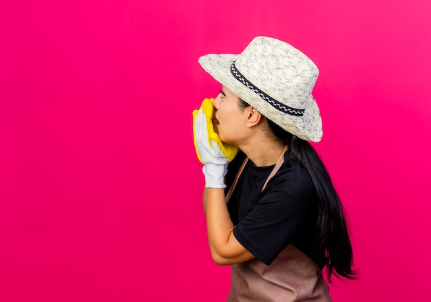 Young beautiful woman gardener in rubber gloves apron and hat calling someone with hands standing over pink wall