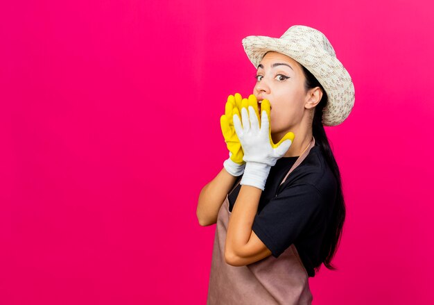 Young beautiful woman gardener in rubber gloves apron and hat covering mouth with hands being surprised standing over pink wall