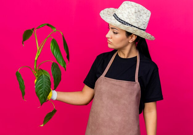 Молодая красивая женщина-садовник в фартуке резиновых перчаток и шляпе, держащая растение, смотрит на него с серьезным лицом