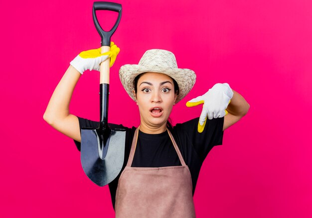 Young beautiful woman gardener in rubber gloves apron and hat holding shovel pointing with index finger down being surprised 