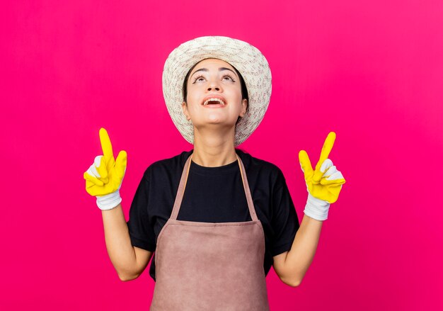 Giardiniere di giovane bella donna in grembiule di guanti di gomma e cappello che punta con il dito indice fino a sorridere