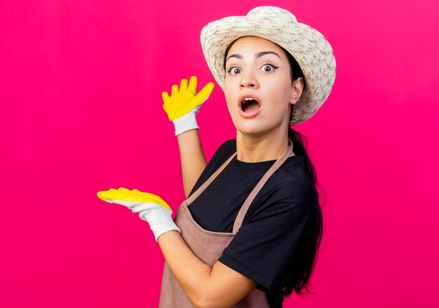 Giardiniere della giovane bella donna in grembiule e cappello dei guanti di gomma che presentano qualcosa con le mani che sono sorpresi e stupiti