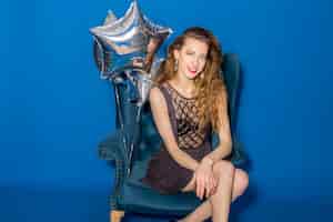 Free photo young beautiful woman in grey dress sitting on a blue armchair with silver balloons