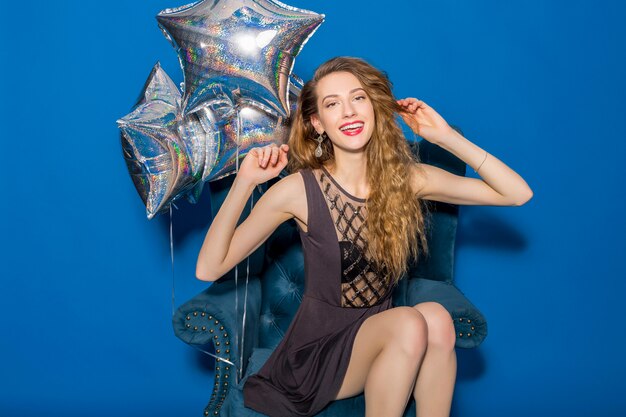 Young beautiful woman in grey dress sitting on a blue armchair with silver balloons