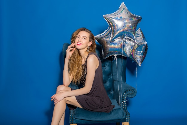 Young beautiful woman in grey dress sitting on a blue armchair with silver balloons