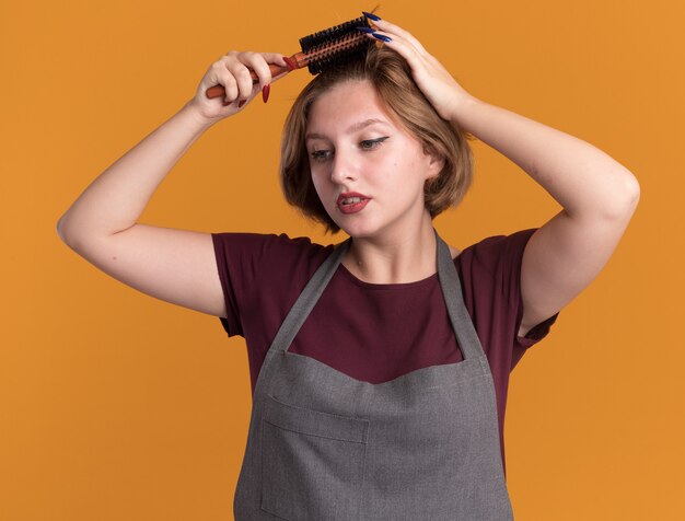 Young beautiful woman hairdresser in apron combing her hair with brush looking aside with serious face standing over orange wall