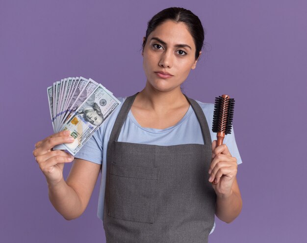 Young beautiful woman hairdresser in apron holding cash and hair brush looking at front with serious face standing over purple wall