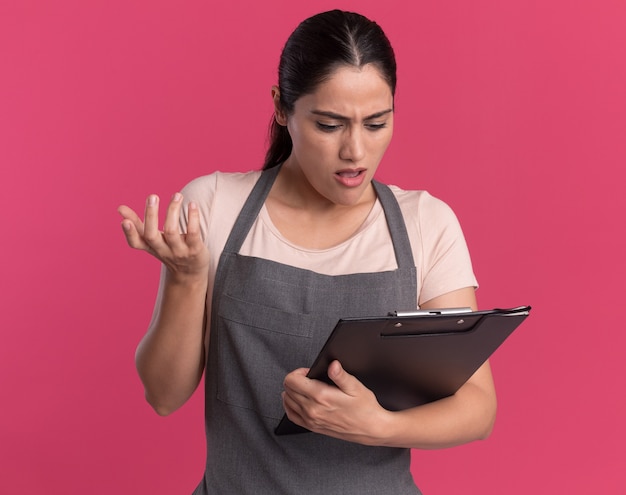 Young beautiful woman hairdresser in apron holding clipboard looking at it being confused and displeased standing over pink wall