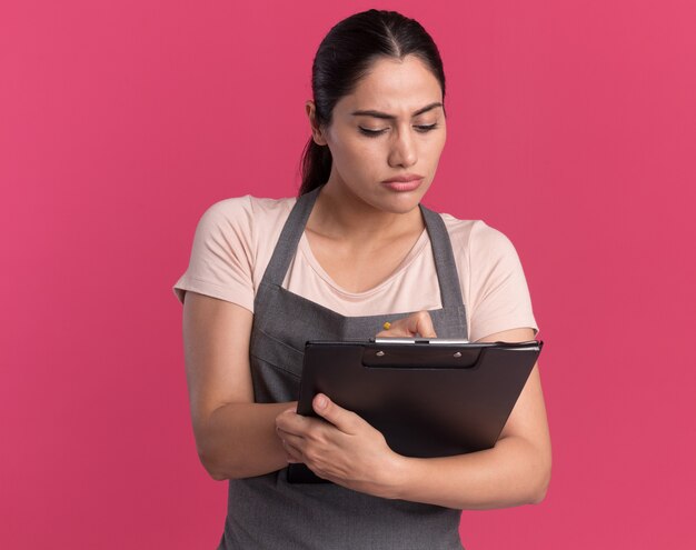 Young beautiful woman hairdresser in apron holding clipboard looking at it with serious face writing something standing over pink wall