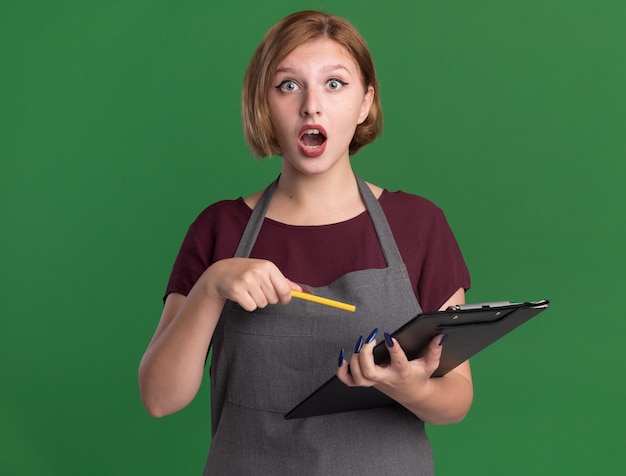 Young beautiful woman hairdresser in apron holding clipboard and pencil looking at front amazed and surprised standing over green wall