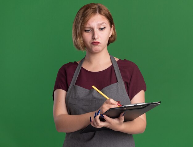 Young beautiful woman hairdresser in apron holding clipboard writing with pencil looking confused and very anxious standing over green wall