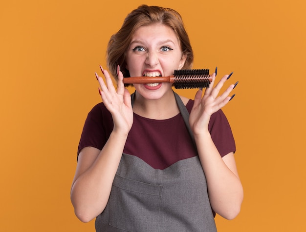 Young beautiful woman hairdresser in apron holding hair brush in her mouth looking at front emotional and excited standing over orange wall