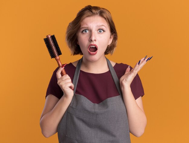 オレンジ色の壁の上に立って驚いて驚いた正面を見てヘアブラシを保持しているエプロンの若い美しい女性の美容師