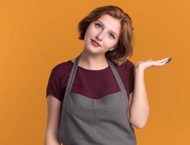 Young beautiful woman hairdresser in apron holding hair brush looking at front with smile on face presenting copy space with arm of her hand standing over orange wall