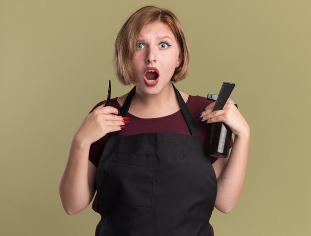 緑の壁の上に立って驚いた正面を見てヘアクリップスプレーボトルと櫛を保持しているエプロンの若い美しい女性の美容師