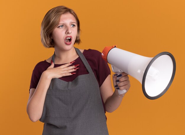 Young beautiful woman hairdresser in apron holding megaphone looking aside confused and surprised standing over orange wall