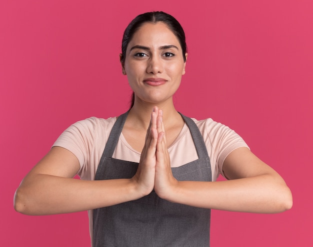 Young beautiful woman hairdresser in apron holding palms together standing over pink wall