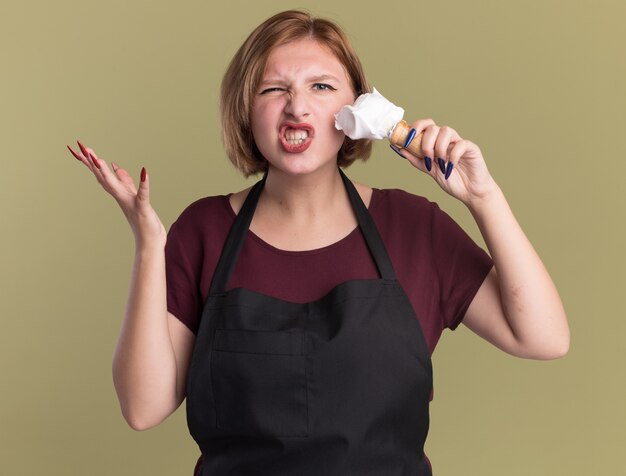 Young beautiful woman hairdresser in apron holding shaving brush with shaving foam looking confused and displeased putting foam on her face standing over green wall