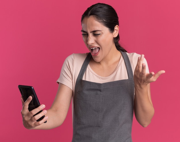 Young beautiful woman hairdresser in apron holding smartphone looking at it being confused and disappointed standing over pink wall