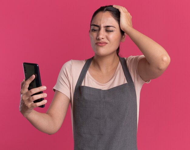 Молодая красивая женщина-парикмахер в фартуке держит смартфон, смущенно и удивленно глядя на него, стоя над розовой стеной