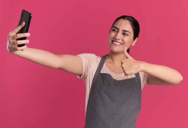 Young beautiful woman hairdresser in apron holding smartphone looking at it doing selfie showing thumbs up smiling standing over pink wall