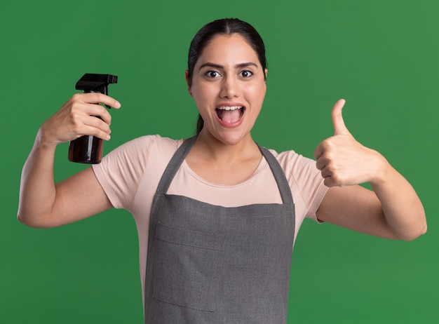 Giovane bella donna parrucchiere in grembiule che tiene la bottiglia spray guardando la parte anteriore sorridente che mostra allegramente i pollici in su in piedi sopra la parete verde