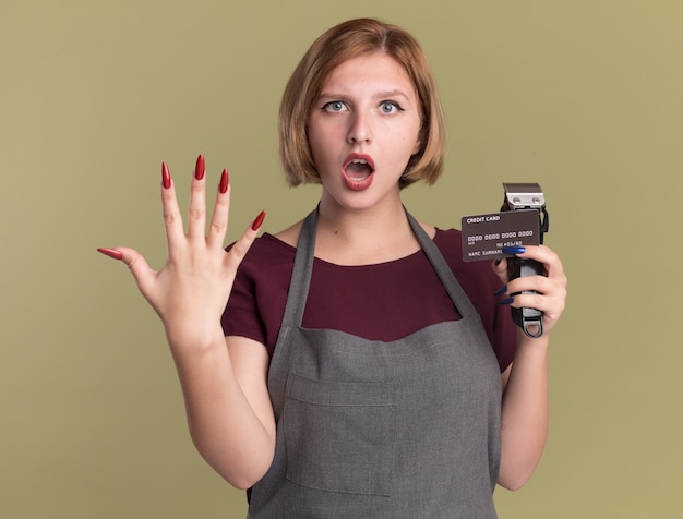 Free photo young beautiful woman hairdresser in apron holding trimmer and credit card looking at front amazed and surprised showing number five standing over green wall