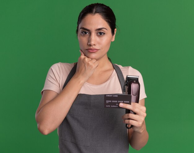 Молодая красивая женщина-парикмахер в фартуке держит триммер и кредитную карту, глядя вперед с рукой на подбородке с уверенным выражением лица, стоя над зеленой стеной