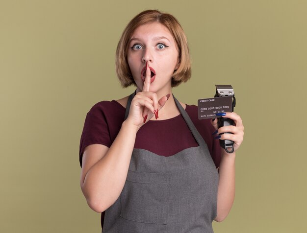 Young beautiful woman hairdresser in apron holding trimmer and credit card making silence gesture with finger on lips looking surprised standing over green wall