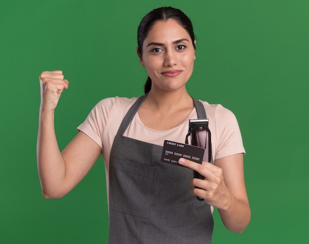 Young beautiful woman hairdresser in apron holding trimmer and credit card raising fist like a winner with confident expression standing over green wall