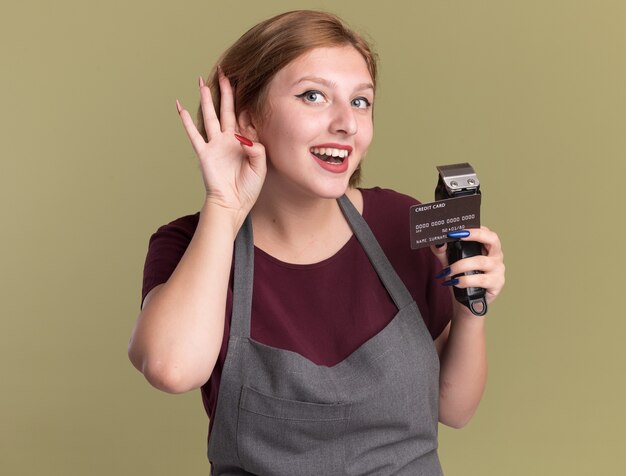 Young beautiful woman hairdresser in apron holding trimmer and credit card with hand over ear trying to listen to gossips standing over green wall