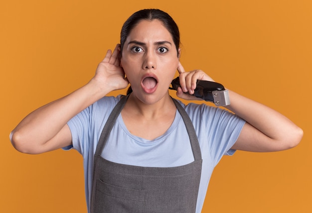 Young beautiful woman hairdresser in apron holding trimmer holding hand over her ear looking surprised standing over orange wall