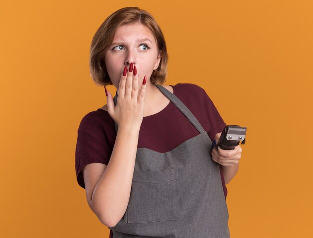 Young beautiful woman hairdresser in apron holding trimmer looking aside being shocked covering mouth with hand standing over orange wall