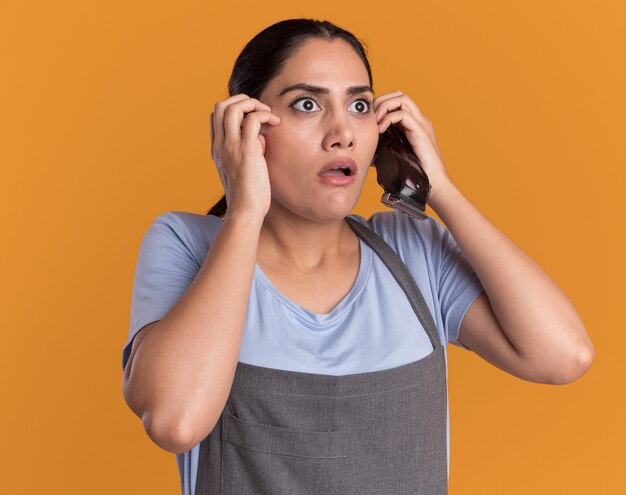 Young beautiful woman hairdresser in apron holding trimmer looking aside scared and worried over orange wall