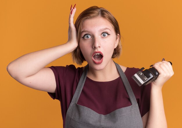 Young beautiful woman hairdresser in apron holding trimmer looking at front surprised standing over orange wall