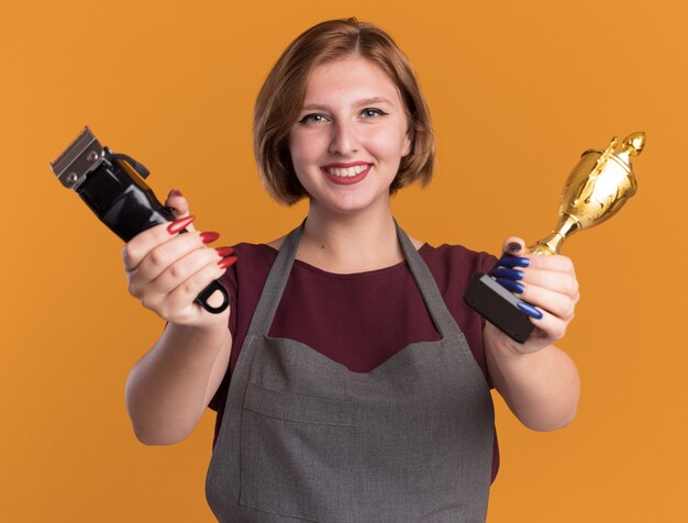 Young beautiful woman hairdresser in apron holding trimmer machine and gold trophy looking at front happy and positive smiling standing over orange wall