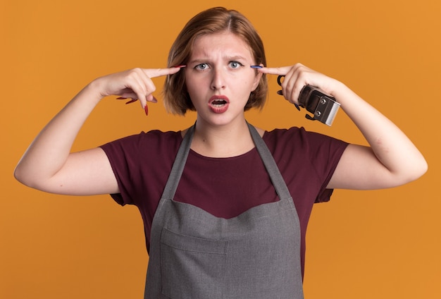 Young beautiful woman hairdresser in apron holding trimmer pointing at her temples looking confused standing over orange wall